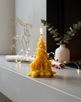 100% beeswax Yellow tree-shaped candle on a mantelpiece with decorative items. pine tree beeswax candle with nativity scene catholic christmas gifts