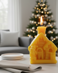 Yellow house-shaped beeswax candle with a flame on a table in a living room with a Christmas tree in the background.