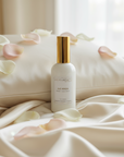 Room spray bottle with gold cap on a white pillow with pink petals. Ave Maria Scent.