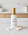 Room and linen spray bottle with gold cap on a marble surface with pink petals, blurred kitchen background.