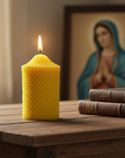 Yellow beeswax candle burning on a wooden surface with a bible and a religious painting of Virgin Mary in the background.