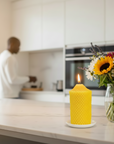 Pure beeswax Yellow candle on a kitchen counter with a person cooking in the background and flowers