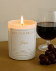 Candle labeled "Jesus" next to a glass of red wine and grapes on a wooden surface, in merlot scent.