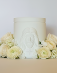 White ceramic jar with embossed face design on a beige surface with flowers