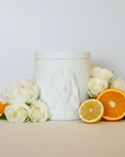 White ceramic jar with embossed face design on a beige surface with flowers and citrus fruits
