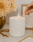 White Embossed Candle with the face of Jesus being lit on a table with flowers, rosary, and card in the background