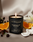 Catholic prayer Candle in a black container with a label that says "It is finished" on a silk fabric background with oranges, coconuts, and flowers representing the scent.