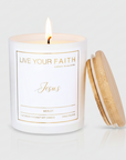 Hand-poured white glass jar candle labeled Jesus Merlot with a burning wick and a bamboo lid leaning against the side.