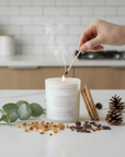 Catholic Prayer Candle being lit with a match, surrounded by eucalyptus leaves and pine cones on a kitchen counter.