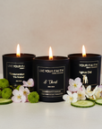 Three lit candles in black containers with 'Live Your Faith' text, surrounded by flowers and cucumbers on a light background.