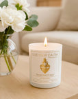 A minimalist lifestyle photograph of the lit white 'Sacred Heart' devotional candle on a light wooden coffee table in a serene living room. Fresh white roses in a glass vase are subtly positioned in the blurred background.