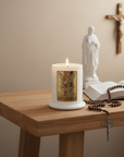 Candle with St. Michael emblem on a wooden table, surrounded by a rosary, open book, and statue.