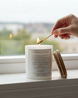 St. Theresa of Avila Candle being lit with a match on a windowsill with a scenic view outside.