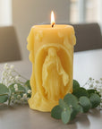 Beeswax candle with a carved design of a figure of virgin Mary on a white background with greenery.