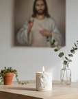 White candle with Pieta design on a table with plants and a blurred portrait in the background