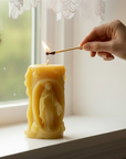 Hand lighting a beeswax candle shaped like Virgin Mary with a window and flowers in the background