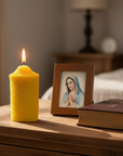 Beeswax Yellow candle, framed picture of Virgin Mary, and a bible on a wooden surface with a blurred bedroom background