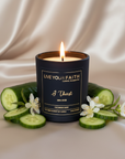 Candle with 'Live Your Faith' branding surrounded by cucumber slices and flowers on a beige fabric background