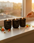 Three black candles on a windowsill with a hand lighting one of them, surrounded by flowers.