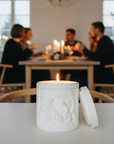 White embossed nativity candle with a burning flame on a table, with a blurred background of people sitting at a table.