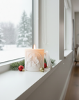 Candle on a windowsill with Christmas decorations and a snowy view outside.