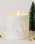 White candle with nativity scene design in a decorative setting with small trees and lights.