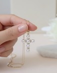 Hand holding a delicate necklace with a cross pendant and pearls against a soft background.