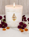 A closeup of the lit white glass 'Sacred Heart' prayer candle, positioned among a composition of golden raw amber pieces, a fresh citrus fruit, a red rose, and small jasmine blossoms to illustrate the golden amber scent profile.