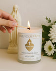 A person’s hand using a long wooden match to light the wick of the 'Sacred Heart' scented prayer candle. The candle is surrounded by a blurry statue of the Blessed Mother and white flowers.
