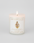 The "Sacred Heart of Jesus'" 7oz scented prayer candle by Live Your Faith Catholic. It is a lit, white matte glass jar with a white paper label and a gold-stamped Sacred Heart graphic, centered against a seamless warm grey studio background.