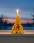 Yellow tree-shaped candle with a lit wick on a windowsill during sunset. pine tree beeswax candle with nativity scene catholic christmas gifts