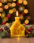 Yellow house-shaped beeswax candle with a lit flame, surrounded by Christmas decorations on a wooden surface with blurred lights in the background.