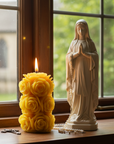 Statue of a religious figure next to a yellow rose-shaped candle by a window.