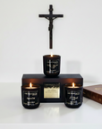 Three lit candles in black containers with visible labels on a white background a bible and a crucifix 