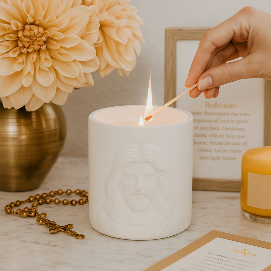 White Embossed Candle with the face of Jesus being lit on a table with flowers, rosary, and card in the background