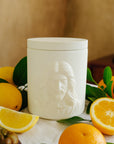 White  jar with the face of Jesus surrounded by fruits and leaves on a table.