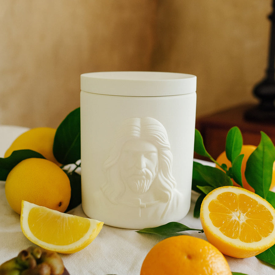 White  jar with the face of Jesus surrounded by fruits and leaves on a table.