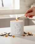 White Pieta embossed candle being lit with a match, surrounded by decorative items on a light surface.