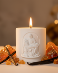 White candle with a decorative design of the Pieta image, surrounded by vanilla beans and amber-colored crystals on a blurred background.