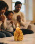 Family praying with a candle shaped like the holy family holding a child on a table in a home setting.