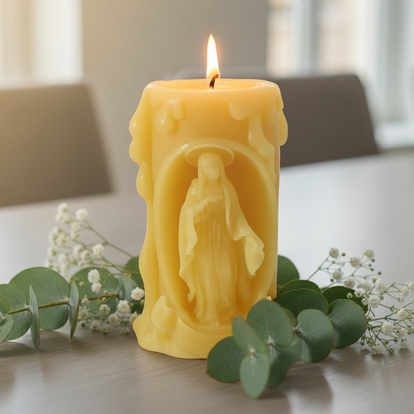 Beeswax candle with a carved design of a figure of virgin Mary on a white background with greenery.