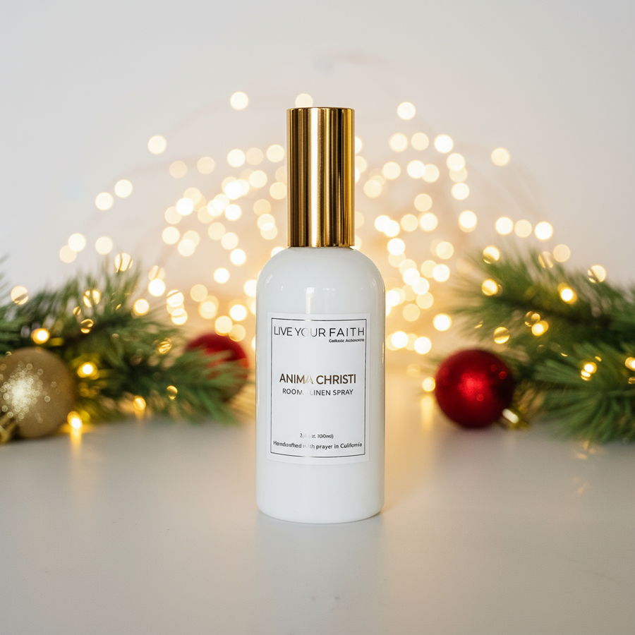 White bottle with gold cap labeled 'Live Your Faith' on a light background with festive decorations. Anima Christi room spray