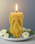Yellow candle with a figurine inside of Virgin Mary on a white plate with flowers, against a blurred background