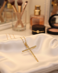 Gold cross necklace on a white pillow with makeup brushes and products in the background