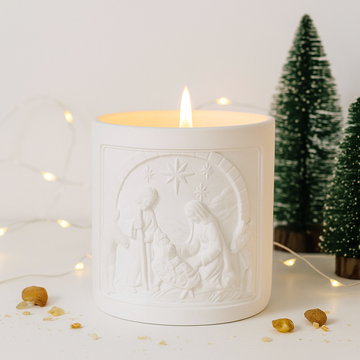 White candle with nativity scene design in a decorative setting with small trees and lights.